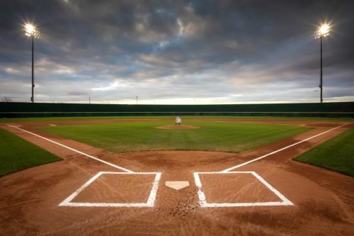 Baseball field at dusk, lit by stadium lights, with a cloudy sky