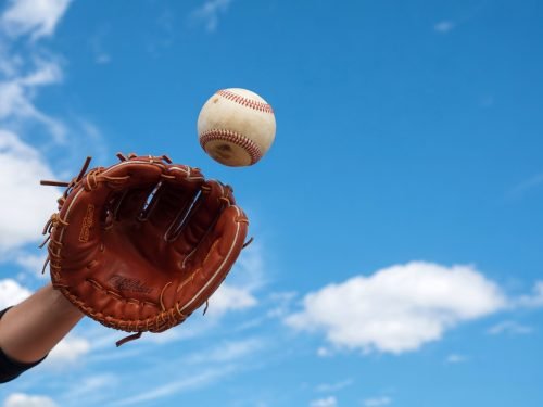 Baseball glove catching a baseball against a bright blue sky with clouds