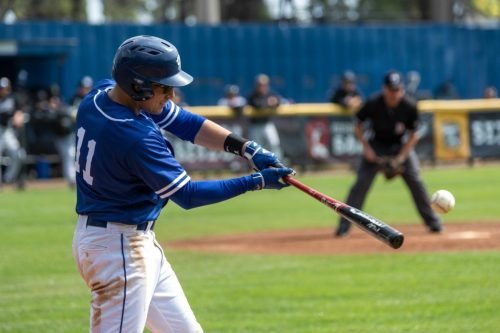 Baseball player in blue uniform swings bat at a pitch, umpire in background