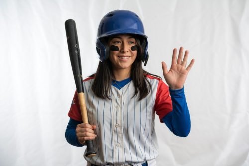 Smiling baseball player in uniform, helmet, holding bat, waving hand